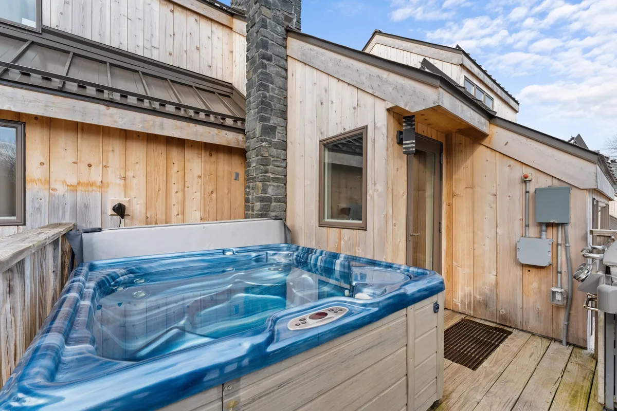 A tranquil exterior setting showcasing a hot tub against a wooden facade and a stone chimney, with snowy surroundings perfect for après-ski relaxation and socializing under the winter sky.