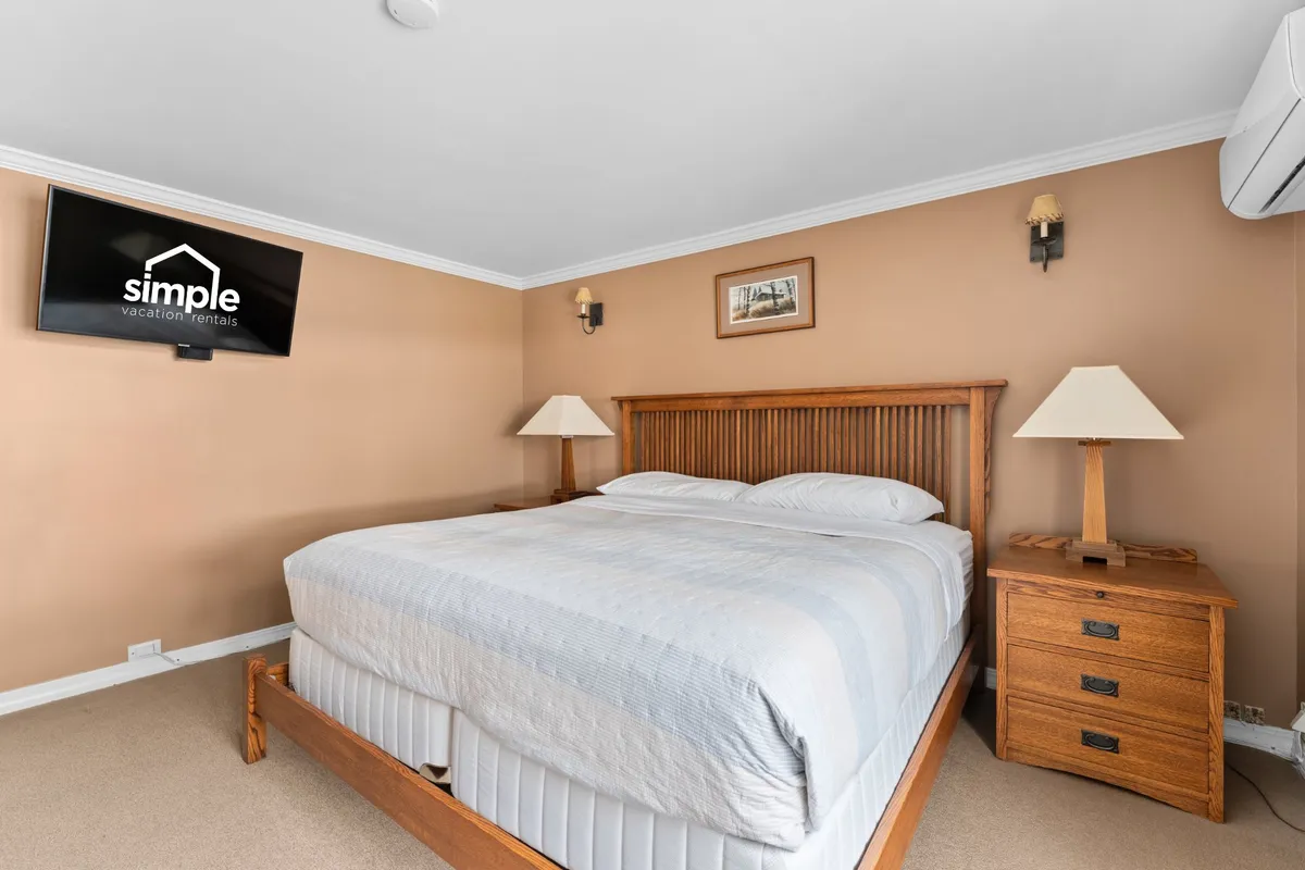 A tranquil bedroom scene featuring a comfortable bed, wall-mounted TV, nightstands, sconce lights, and a framed picture. The atmosphere is restful and well-appointed, inviting guests to relax or enjoy quiet downtime. The setup reflects both comfort and technology, with a TV included for individual entertainment.