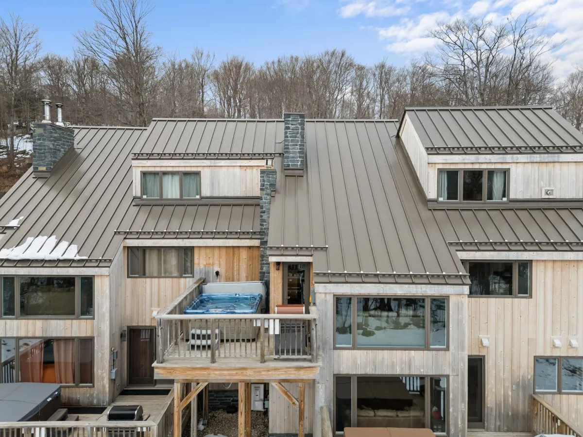 This exterior image features a hot tub situated on a wooden deck, with a view of a modern mountain home. Snow dusts the railing and roof, emphasizing its year-round appeal and the connection to the outdoors. Large windows and a stone chimney add warmth and a sense of shelter.