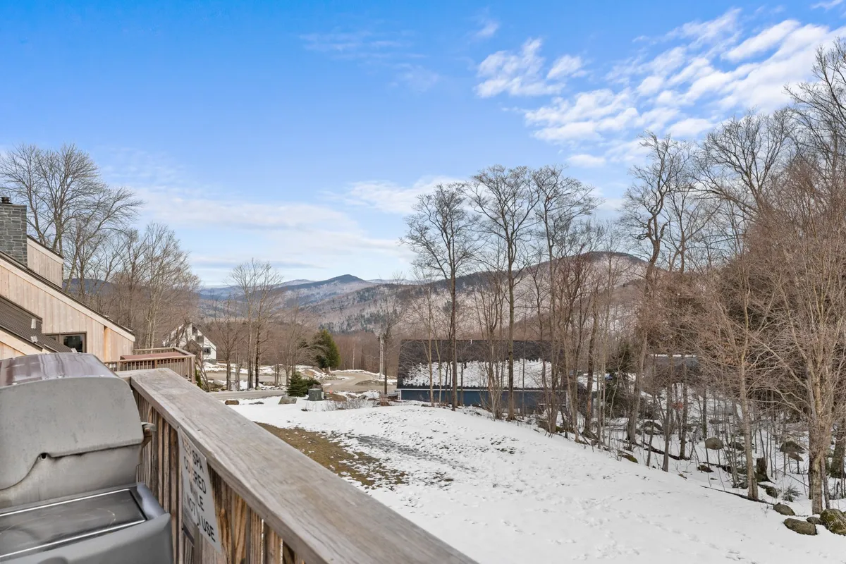 A peaceful view from a deck, looking out over a snow-covered ground, bare trees, and distant mountains. The deck railing hints at private outdoor space perfect for relaxing after a day of adventure, while the mountains and trails invite exploration in every season.