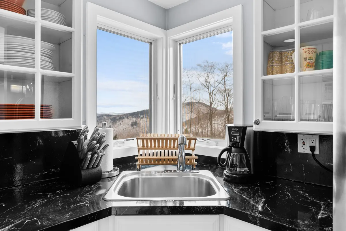 A cozy kitchen corner with a sink, a coffee maker on the counter, a dish rack, and windows that allow natural light to flood in. The setup suggests a welcoming spot for morning routines and meal prep while enjoying scenic views.