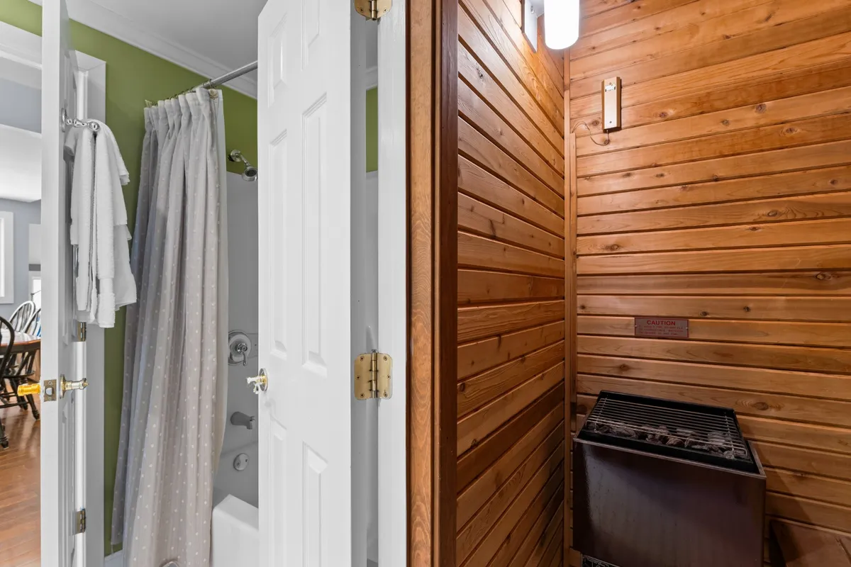 An interior scene featuring a wood-paneled wall, a shower area with a curtain, and a small grill or heating element. The warm wood tones create a sauna-like ambiance, evoking the cedar sauna described in the section.