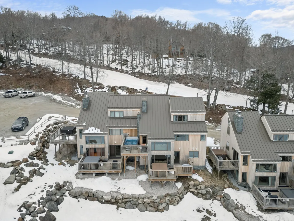 An inviting exterior scene with wooden balconies, a steaming hot tub, and snow-covered ground, set against a backdrop of rock formations and mountains. The image exudes warmth and relaxation after a day on the slopes.