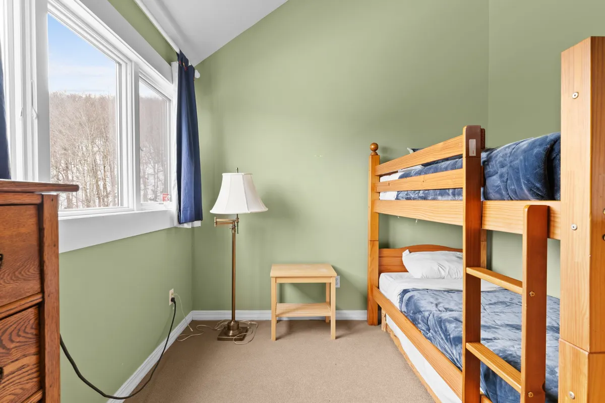 A cozy bedroom with a twin bunk bed, soft lamp lighting, a nightstand, and windows allowing natural light to filter in. The setup is inviting and playful, perfect for children or friends, emphasizing the flexibility of sleeping arrangements and the fun of sleepovers or shared family moments.