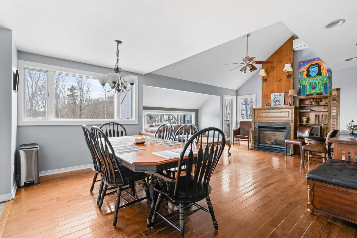 A warm interior view featuring a large dining table set beneath expansive windows, with a cozy fireplace and vibrant artwork on the wall. The space is filled with natural light, accentuating the inviting atmosphere for group meals and gatherings.