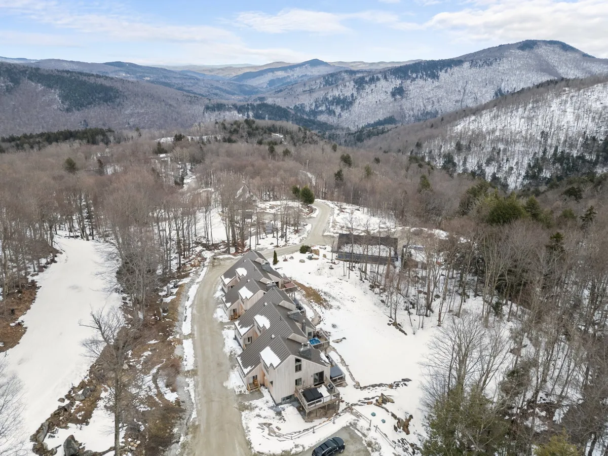 A tranquil, snow-covered landscape dotted with a cluster of houses, winding road, and expansive mountain views. The scene captures the natural beauty and direct access to trails, perfect for various seasonal outdoor activities, with a sense of peaceful adventure.